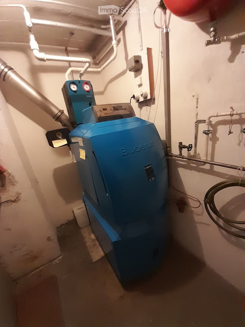 A blue boiler is mounted on the wall, with pipes and wires connected to it. A white container is placed on the floor near the boiler. A red tank is mounted on the ceiling. A wooden plank is placed on the floor.