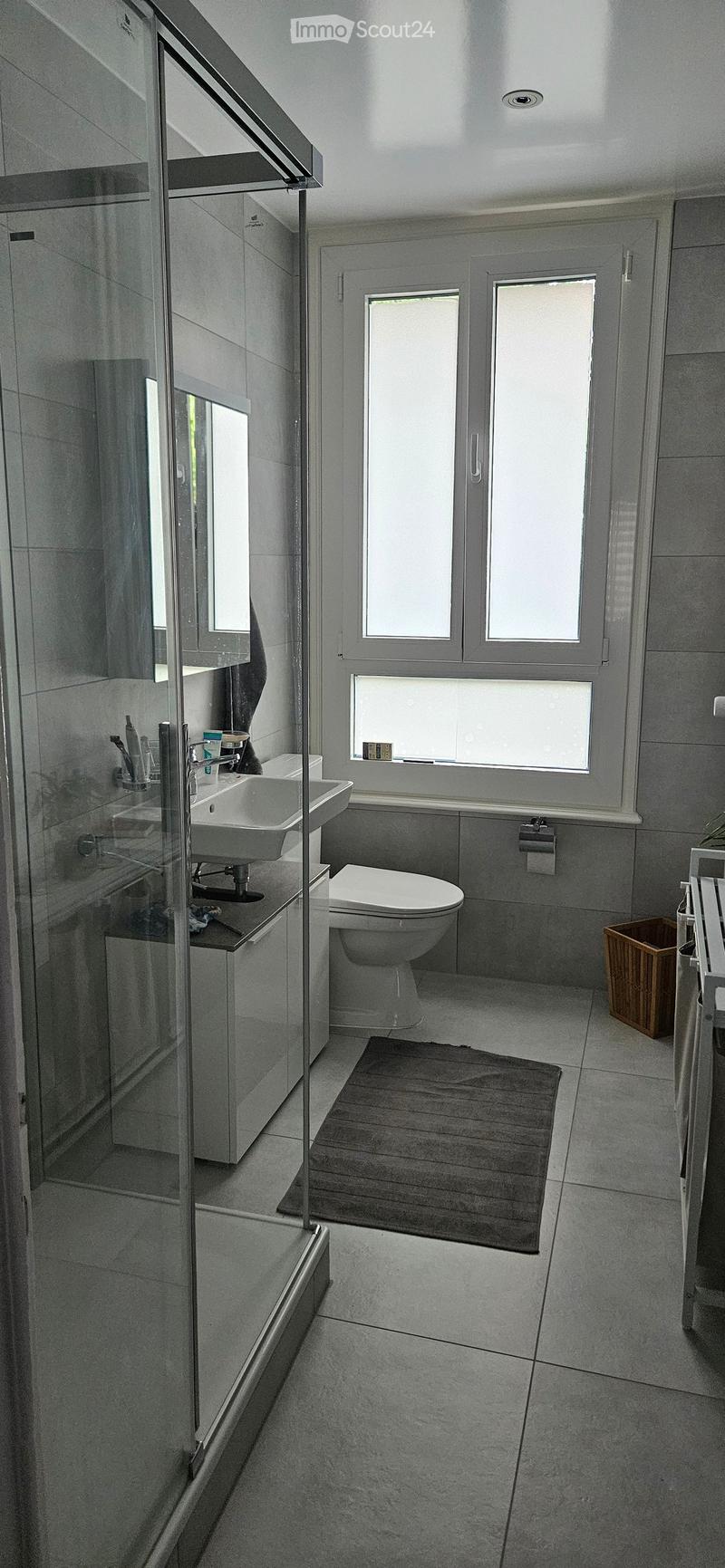Modern bathroom with grey tiles, a large window, a shower with a glass door, a sink with a mirror, a toilet, and a small rug on the floor.