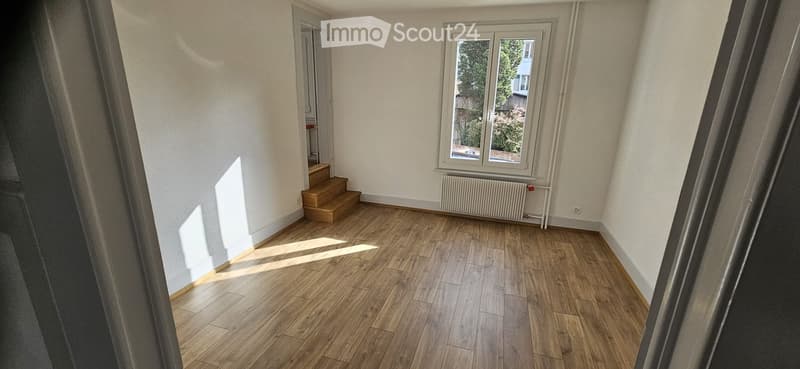 Empty room with wooden flooring, white walls, a window with a view of greenery, a radiator, and a small staircase leading to another area.