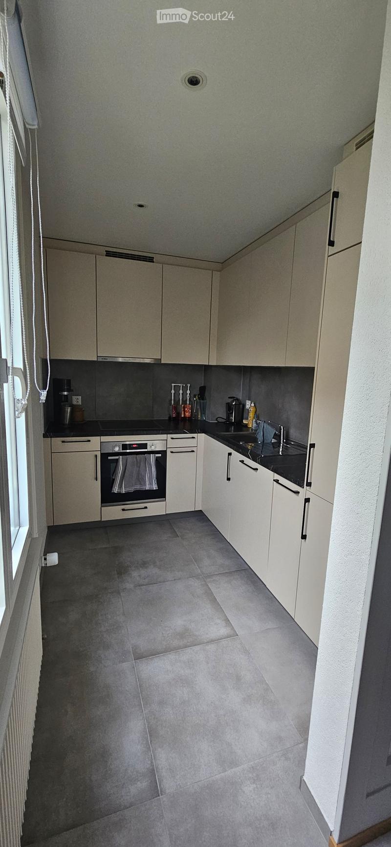 Modern kitchen with light-colored cabinets, a black countertop, and a tiled floor. Features an oven, sink, and various small appliances.