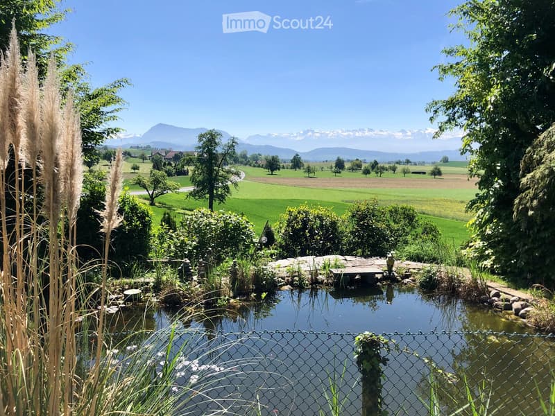 A serene garden with a pond, surrounded by lush greenery, tall grasses, and a view of distant mountains.