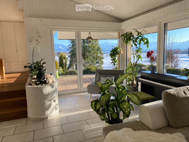 Bright living room with large windows offering a scenic mountain view, featuring a sofa, piano, potted plants, and a decorative buddha statue on a round table.