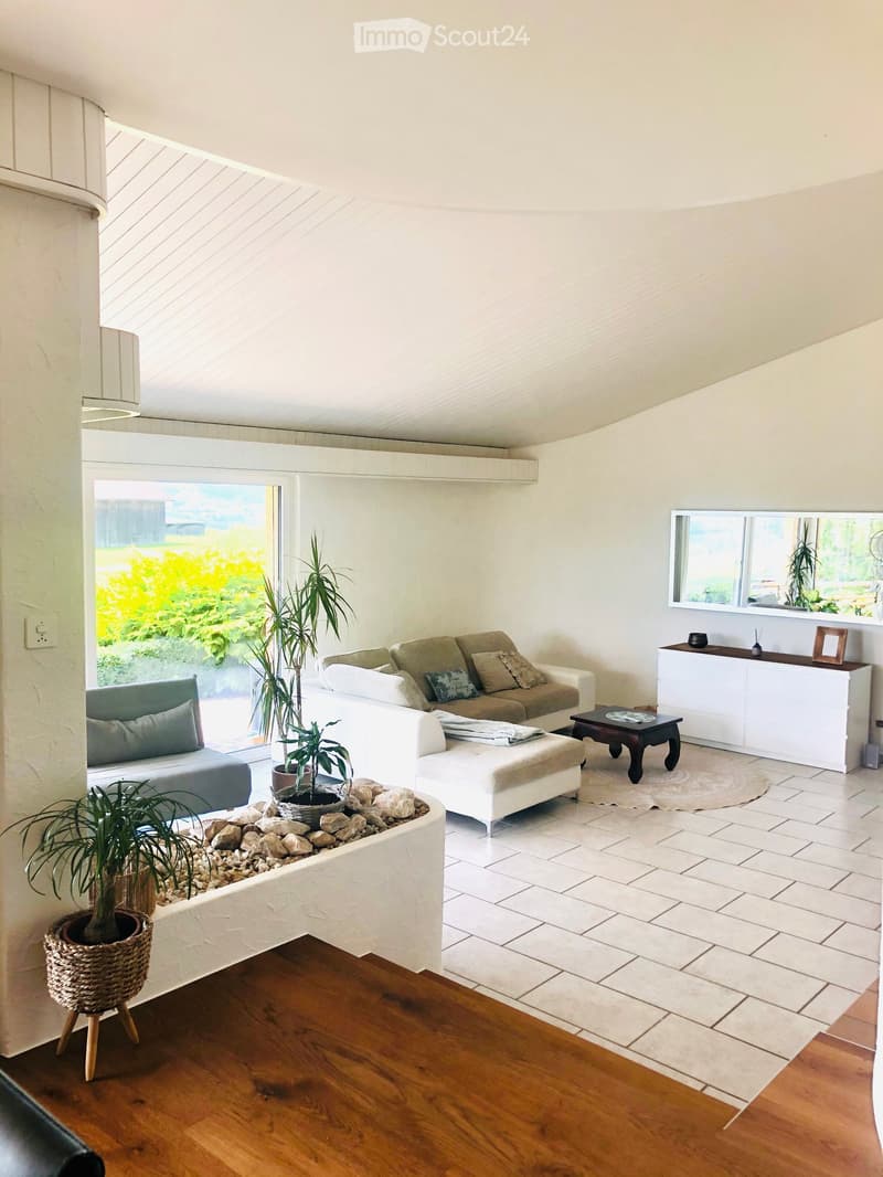 Bright living room with white walls, large windows, and skylight. Features a beige sofa with pillows, white ottoman, coffee table, side table, and a white cabinet with a mirror above it. Potted plants are placed around the room, and the flooring is a mix