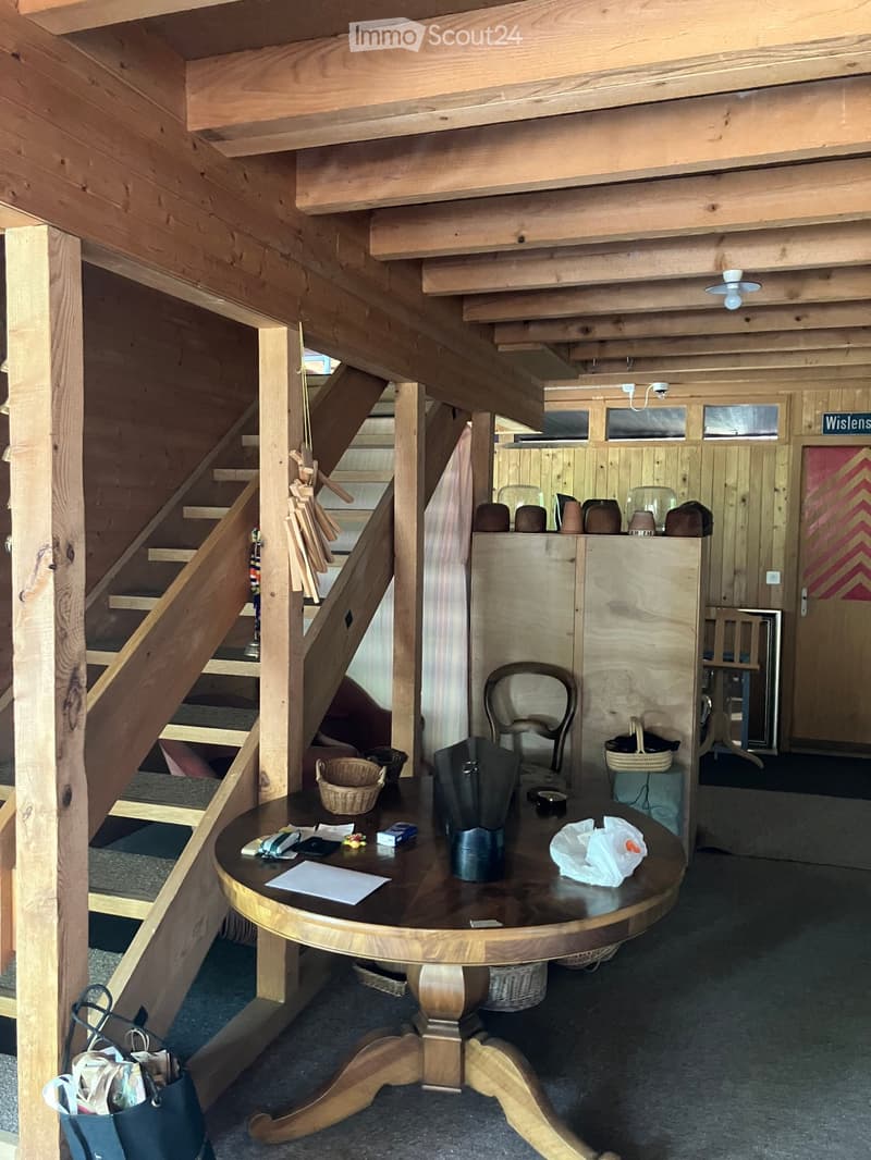 Wooden interior with a round wooden table, a chair, a cabinet, and baskets. Stairs lead upwards, and a door is visible in the background.