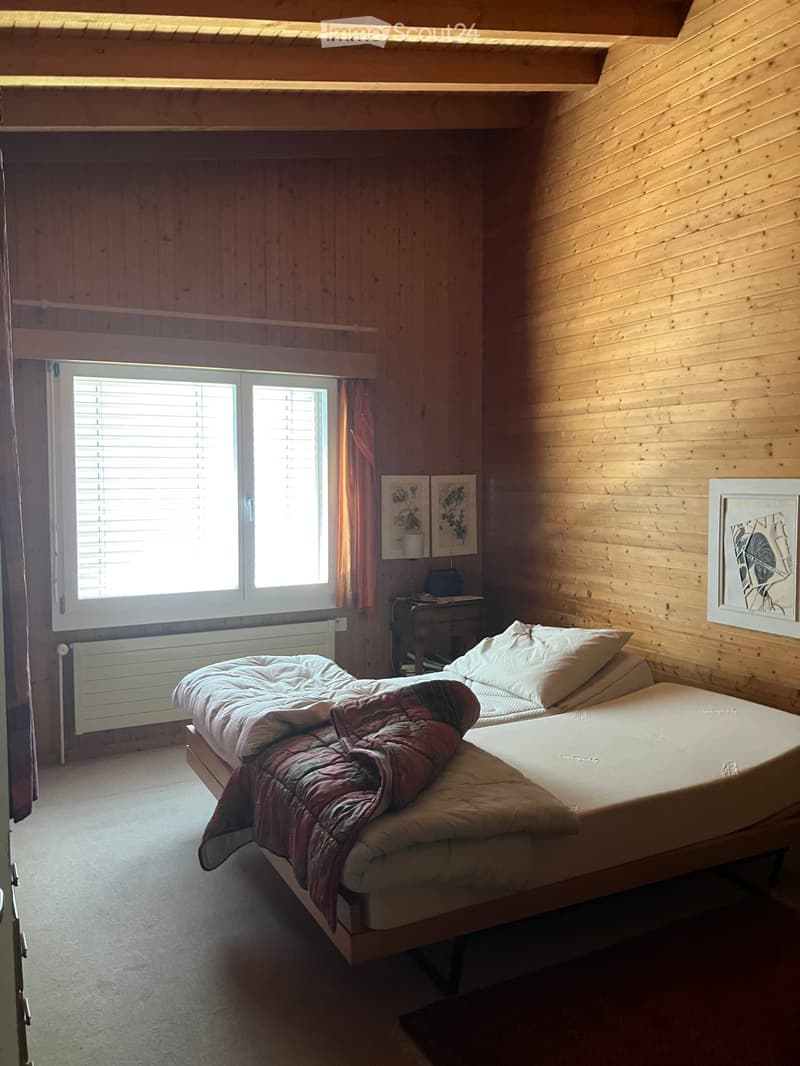 Cozy wooden bedroom with a double bed, nightstand, lamp, and artwork on the walls. Large window with blinds allows natural light to enter.