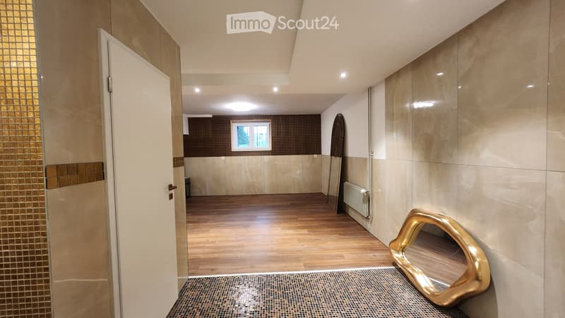 Modern hallway with wooden flooring, beige tiled walls, and recessed lighting. Features a large gold mirror and a small window at the end.