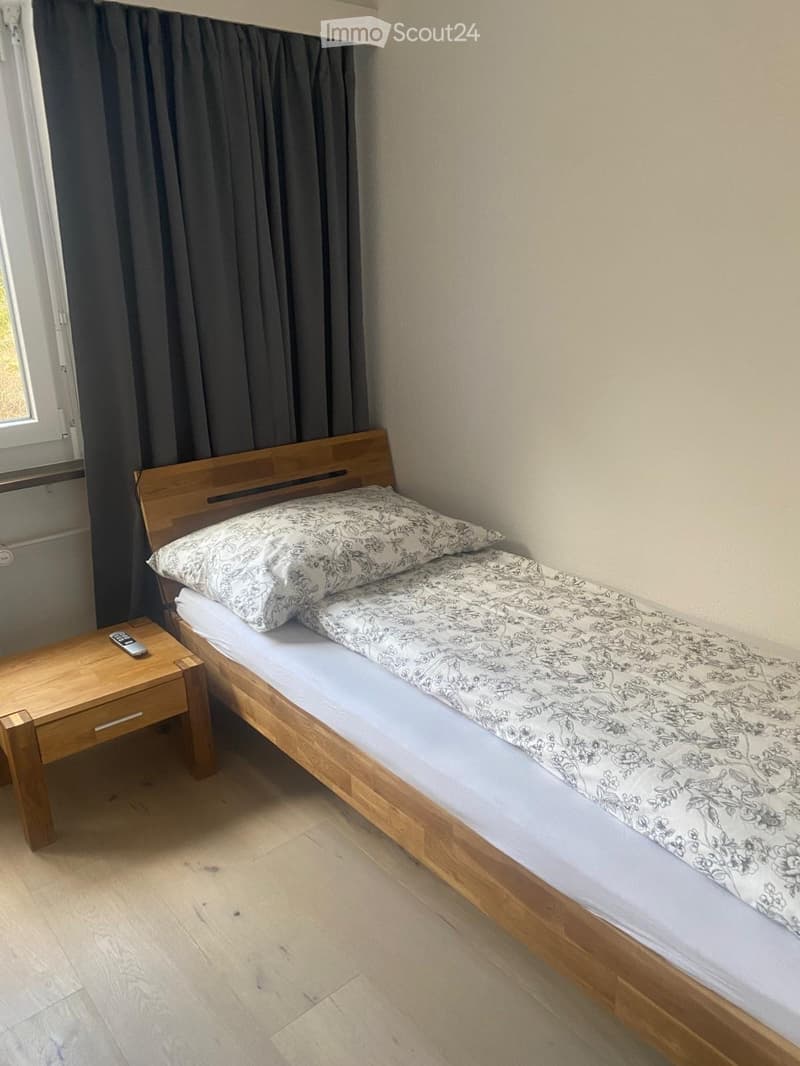 A small bedroom with a single wooden bed, patterned bedding, a wooden nightstand, and a remote control on top.