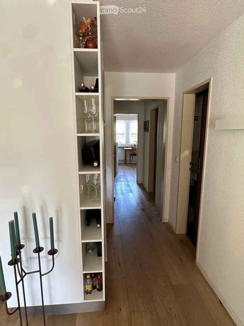A well-lit hallway with wooden flooring, white walls, and a tall white shelving unit on the left side holding wine bottles, glasses, and decorative items. The hallway leads to an open doorway revealing a room with a table and chairs.