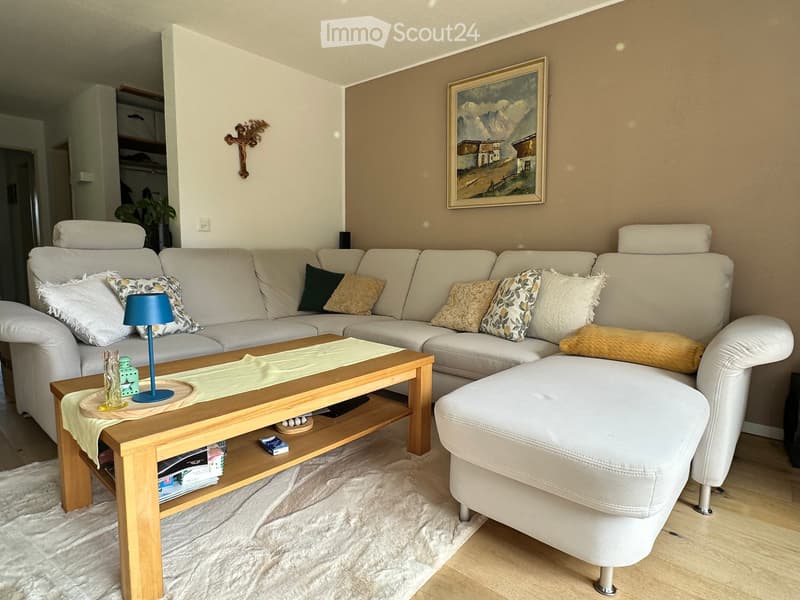 Modern living room with a white sectional sofa, wooden coffee table, ottoman, and decorative pillows. Features a table lamp, picture frame, and a small plant in the corner.