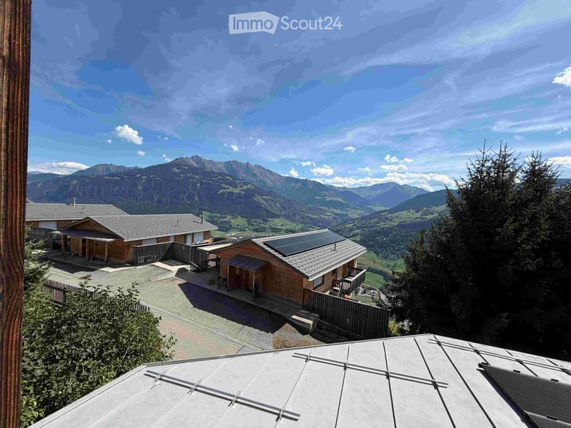 A panoramic view of a house complex with solar panels on the roof, surrounded by lush greenery and a stunning mountain backdrop.