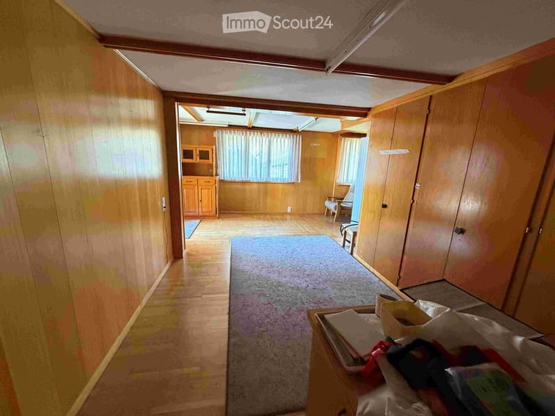 A narrow hallway with wooden walls and carpeted floor, leading to a room with a small table, chair, and window with curtains.