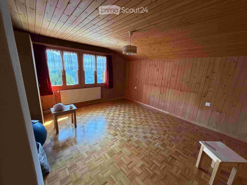 Empty room with wooden walls, wooden floor, and large windows with curtains. A small table and a bench are present, but the room is otherwise unfurnished.