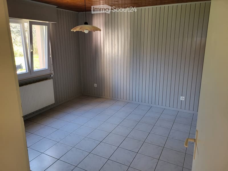 Empty room with tiled floor, wooden panel walls, and a hanging light fixture. Windows and radiator visible.