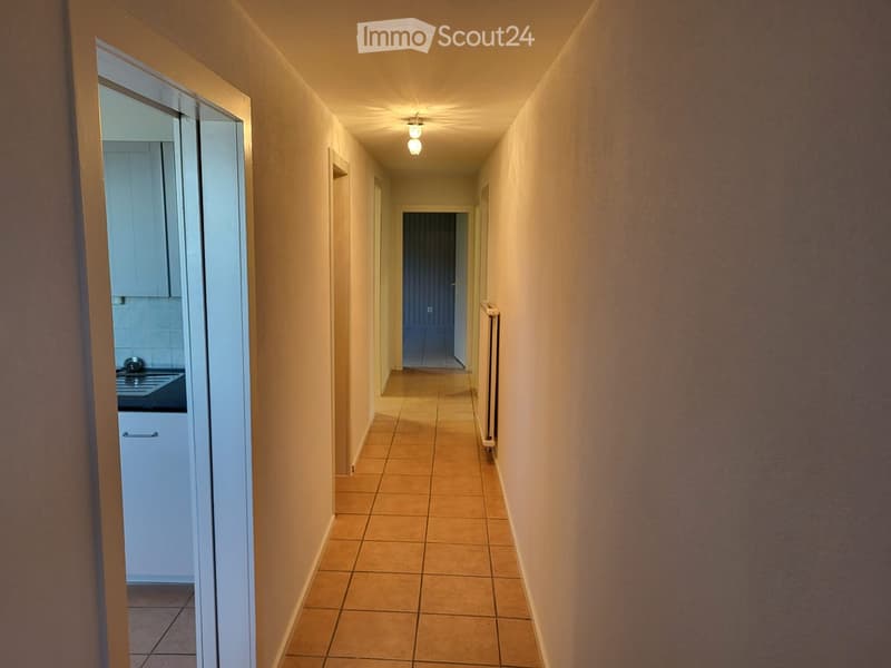 A narrow hallway with beige tiled flooring, light-colored walls, and a ceiling light fixture. An open door on the left leads to a kitchen area with white cabinets and a sink, while a closed door at the end of the hallway is visible.