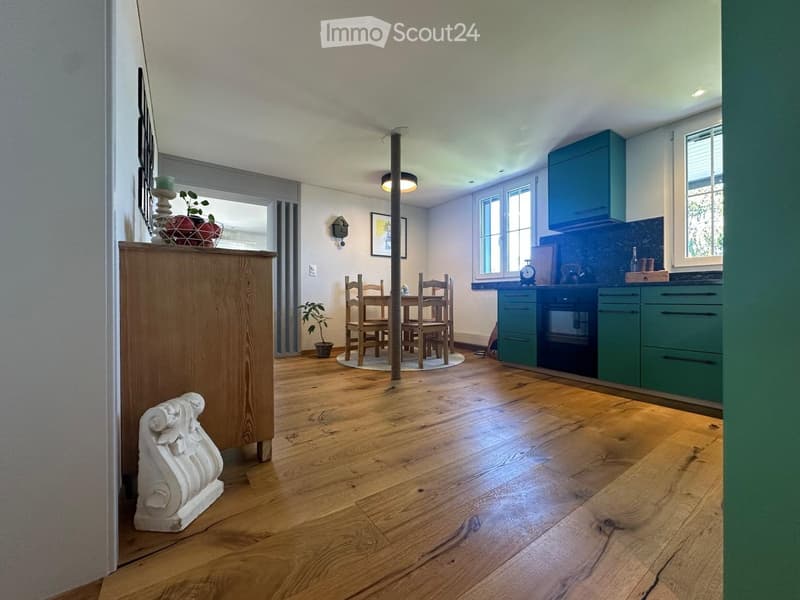 Open-plan kitchen and dining area with wooden flooring, green kitchen cabinets, a dining table with chairs, and decorative items including a vase, candle, and framed picture.
