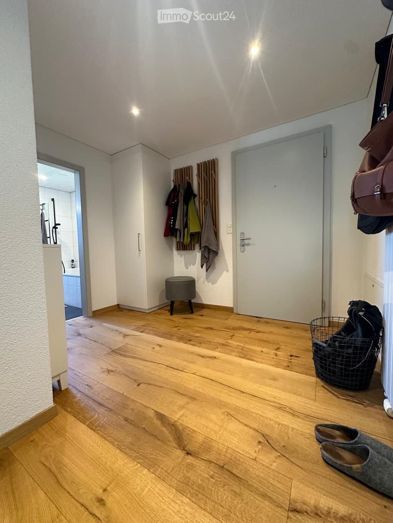 A well-lit hallway with wooden flooring, white walls, and a coat rack holding several jackets and coats. A small round stool is placed near the coat rack, and a wire basket with a backpack is on the floor. A pair of slippers is also visible near the baske