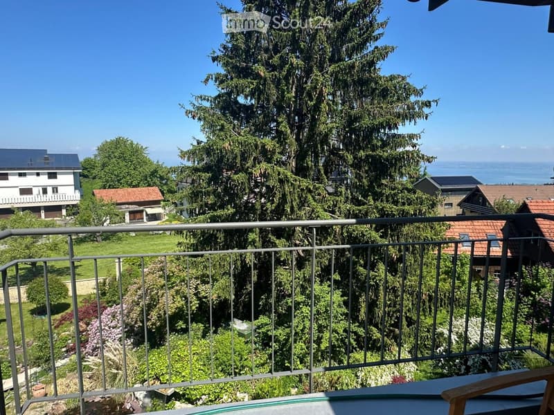 Balcony overlooking a lush garden and residential area with houses and trees, featuring a metal railing and a wooden chair.
