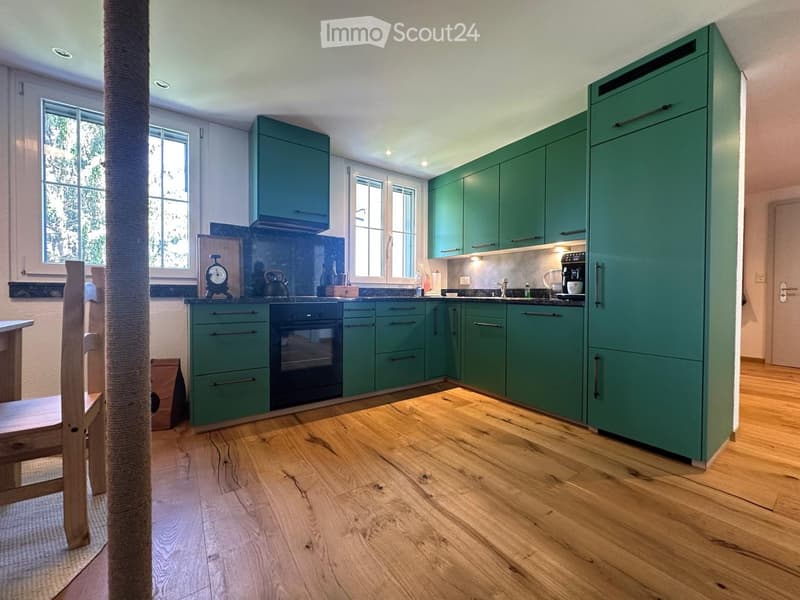 Modern kitchen with green cabinets, wooden flooring, and stainless steel appliances including a refrigerator, oven, and coffee maker. A wooden chair is visible near the window.