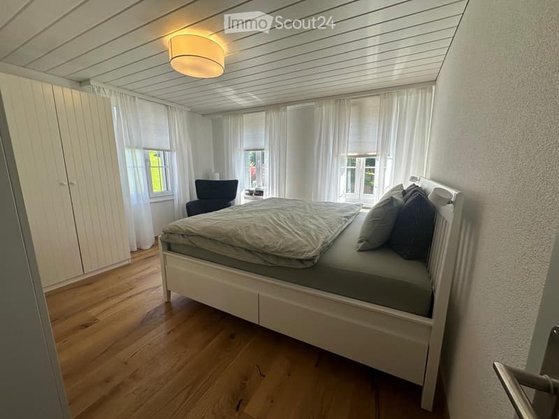 Cozy bedroom with a white bed, grey bedding, two pillows, a black chair, and a white wardrobe. The room features wooden flooring, large windows with white curtains, and a modern ceiling light.