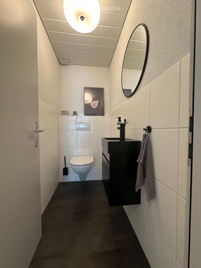 Modern bathroom with white tiled walls, a toilet, a black sink with a modern faucet, a round mirror, a towel, and a picture frame on the wall.