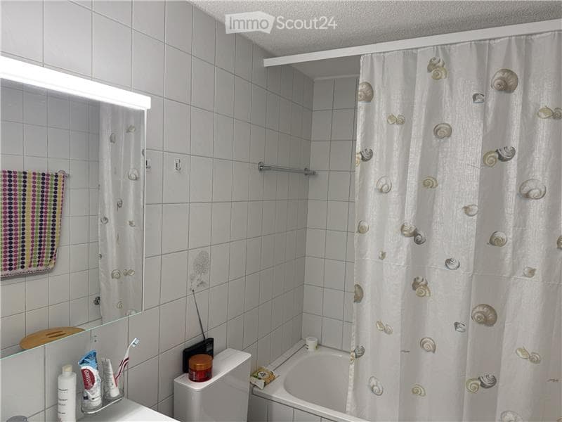 A small bathroom with white tiled walls and floor, featuring a bathtub with a shell-patterned shower curtain, a toilet, a mirror with a light fixture above it, and a towel hanging on the wall. Personal care items are placed on the sink counter.