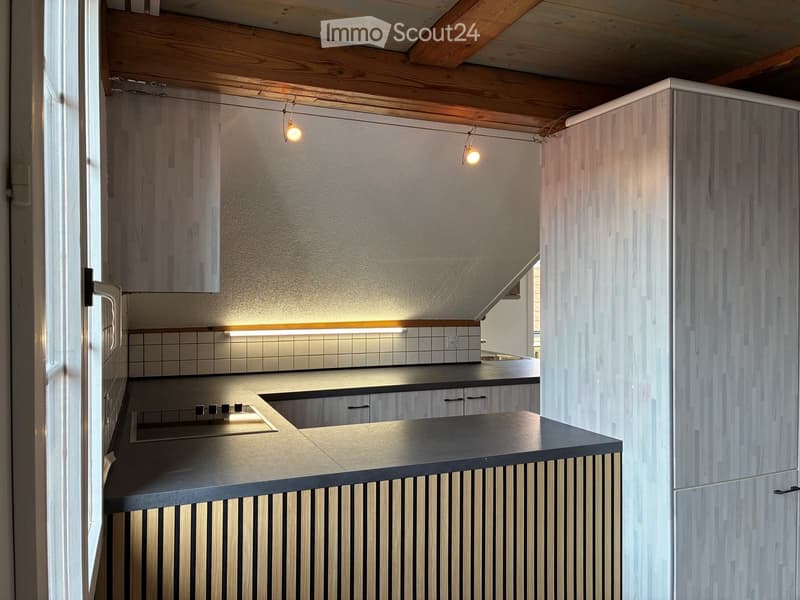 Modern kitchen with sleek wooden cabinets, a black countertop, and built-in appliances. Exposed wooden beams on the ceiling and warm lighting.