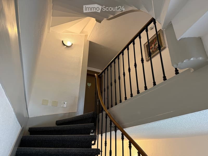 Spiral staircase with black wrought iron railing and wooden handrail, carpeted steps, wall-mounted light fixture, and a framed picture on the wall.