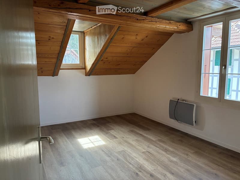 Empty attic room with wooden beams, wooden flooring, and large windows allowing natural light. A small radiator is mounted on one wall.