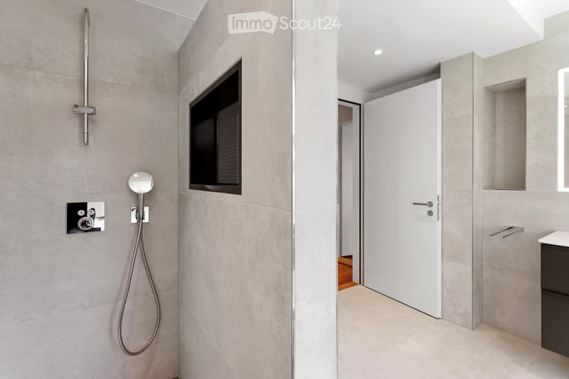 Modern bathroom with grey tiled walls, a sleek showerhead, a built-in mirror, and a dark cabinet.