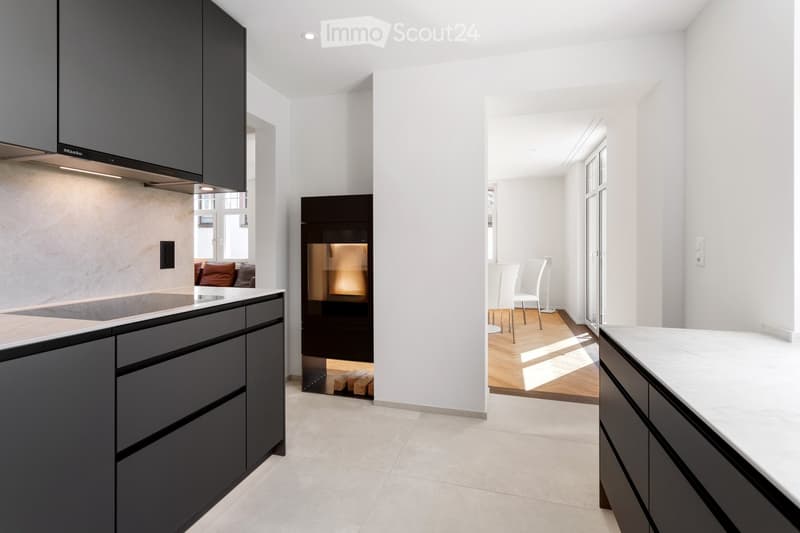 Modern kitchen with dark grey cabinets, marble countertops, and a central island. Adjacent living area features a fireplace, white chairs, and a sofa.