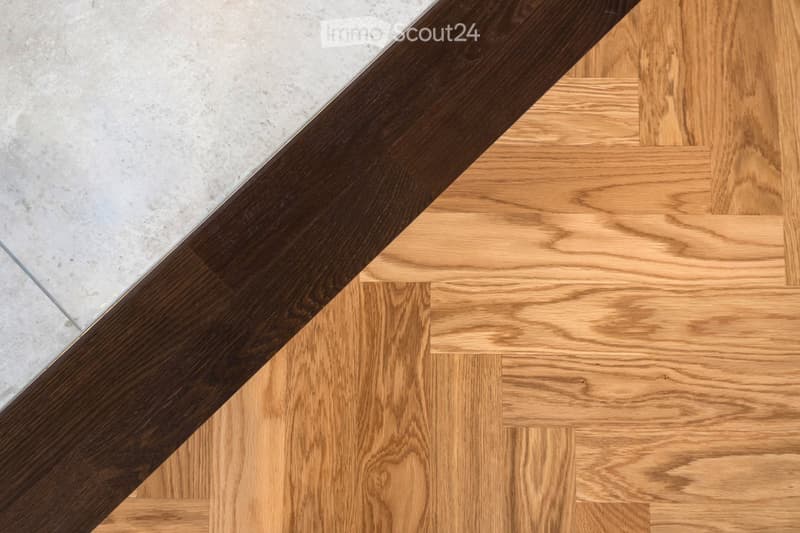 Close-up view of a transition between a polished concrete floor and a light wooden parquet floor, featuring a dark wooden skirting board.