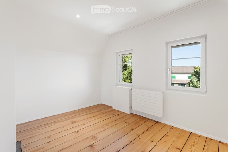 Empty room with white walls, wooden floor, two windows, and white radiators.