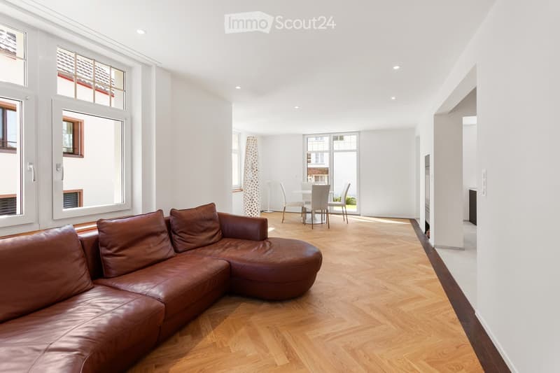 Open-plan living and dining area with light wooden flooring, large windows, a brown leather sofa, a white dining table with chairs, and a decorative floor lamp.