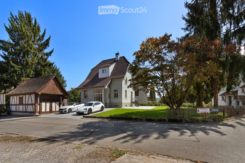 Two-story house with a brown tiled roof, white walls, and large windows. A wooden garage is attached to the left side, and two white sports cars are parked in front. The house is surrounded by trees and a well-maintained lawn.