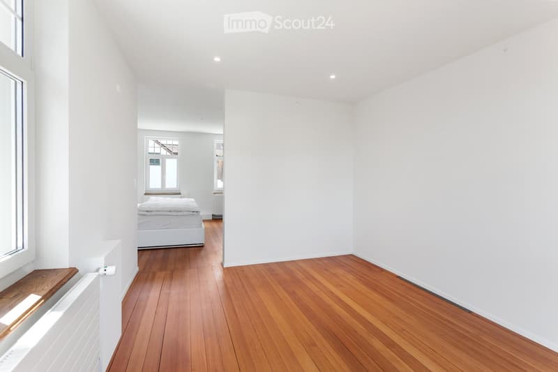 Bright, minimalist bedroom with wooden flooring, white walls, and a single bed visible in the background. The room features a radiator and a window with a wooden bench.