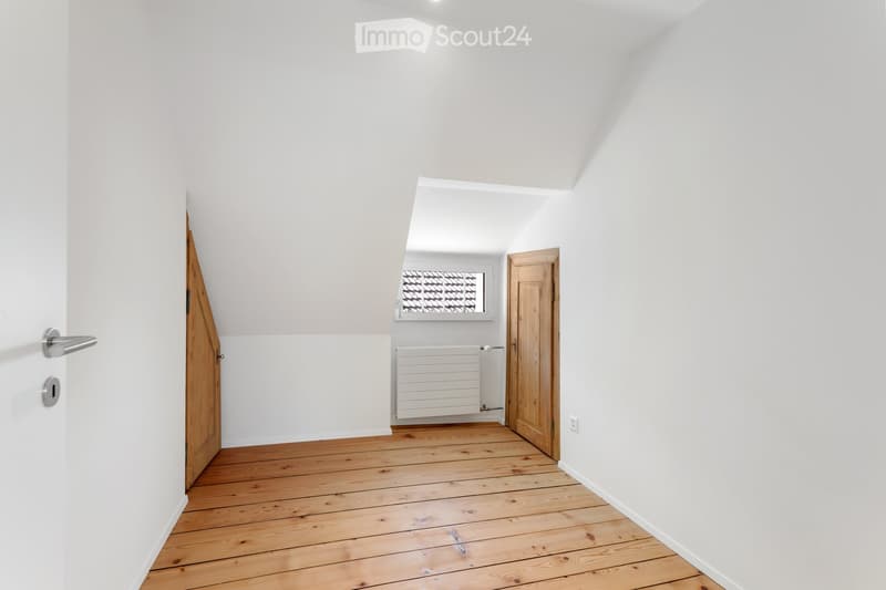 Empty room with wooden flooring, white walls, and two wooden doors. A radiator is visible in the background, and a small window with a decorative grid pattern is present.