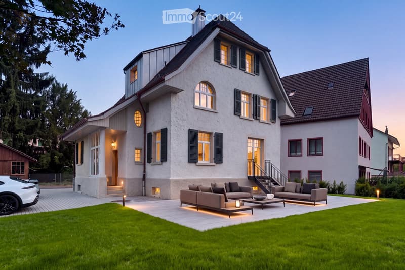 A modern two-story house with white exterior walls, dark green shutters, and multiple windows. The house features a well-maintained lawn, a paved patio area with outdoor seating, and a white car parked nearby.