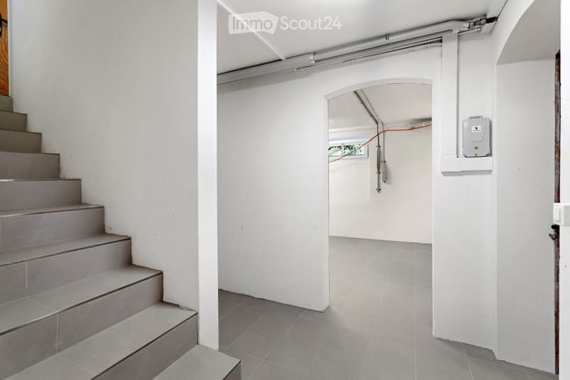 A clean, modern hallway with white walls and grey tiled flooring. A staircase leads upwards on the left, and an open doorway on the right reveals a glimpse of another room with visible electrical equipment.