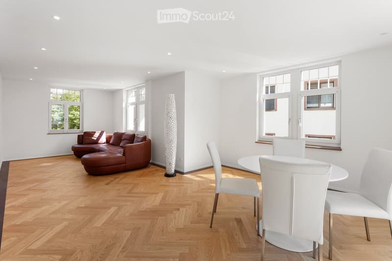 Bright living and dining area with wooden flooring, large windows, a brown leather sofa, a white dining table with chairs, and a decorative floor lamp.