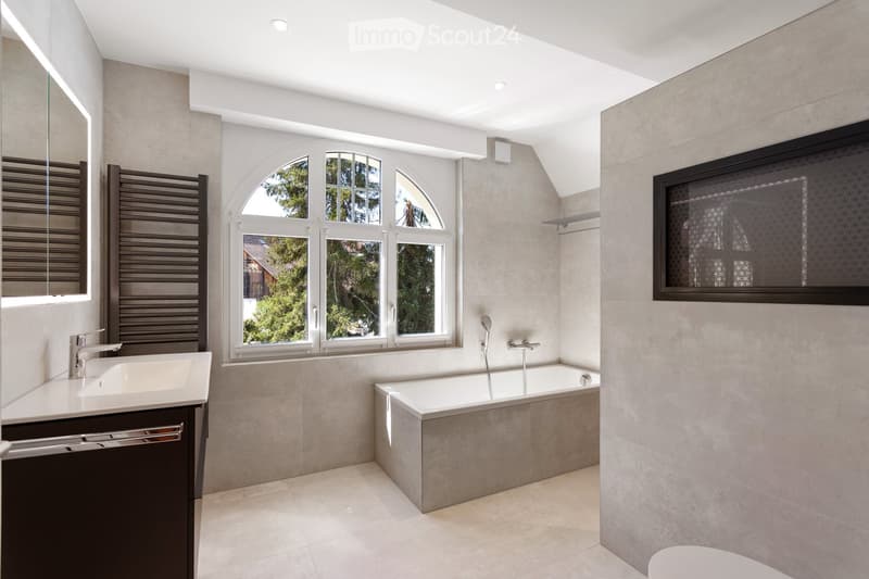 Modern bathroom with a large window, bathtub, sink, and mirror. Features include a radiator, sleek fixtures, and a contemporary design.