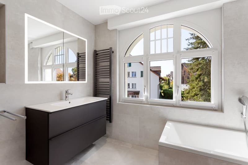 Modern bathroom with a dark vanity, white sink, large mirror, bathtub, and arched windows offering a view of greenery.