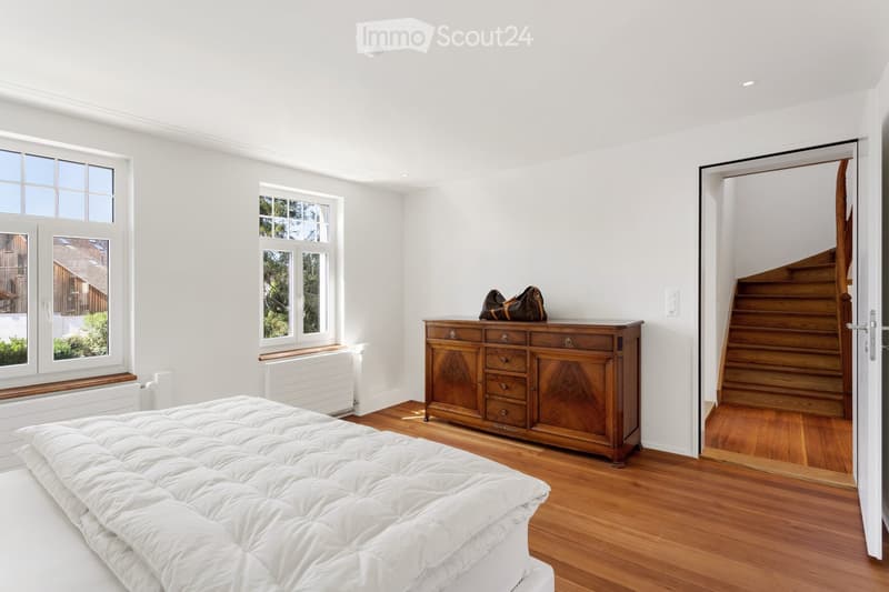 Bright bedroom with wooden flooring, large windows, a neatly made bed, a wooden dresser with drawers, and a handbag on top.