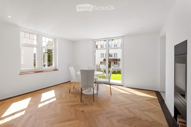 Bright, minimalist dining area with white walls, wooden herringbone flooring, and large windows. A white dining table with four matching chairs is positioned near the window, allowing natural light to fill the space.