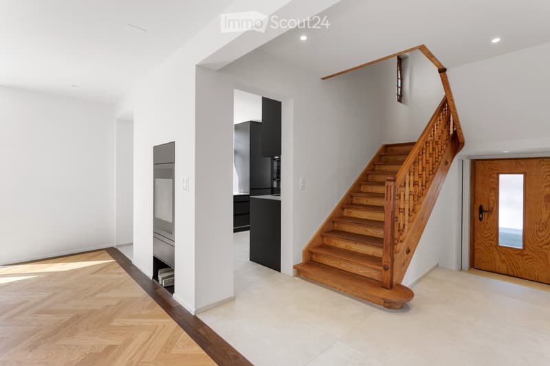 Bright, modern hallway with wooden staircase, white walls, and polished wooden floors. Features a built-in cabinet and a door leading to another room.