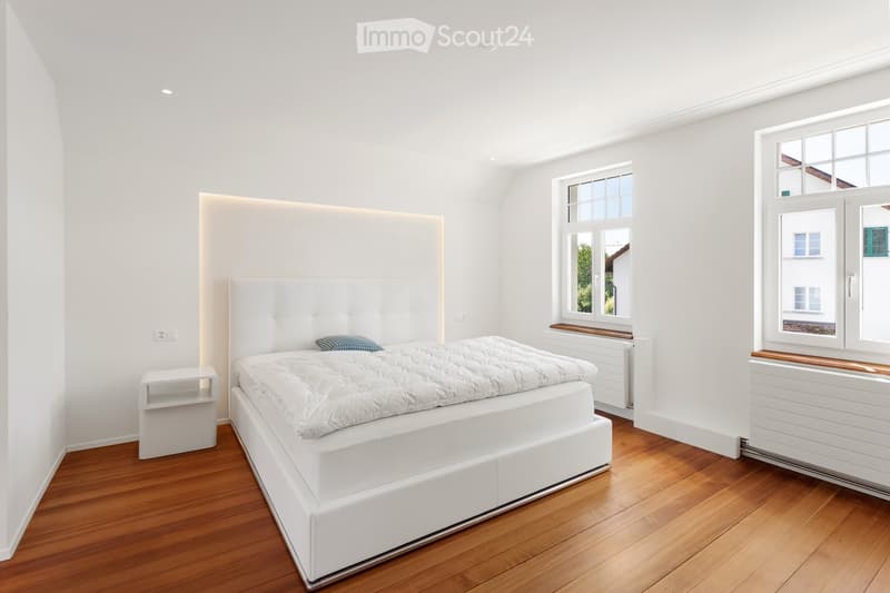 Modern bedroom with white walls, wooden floor, large bed with white headboard, nightstand, and windows with wooden sills.