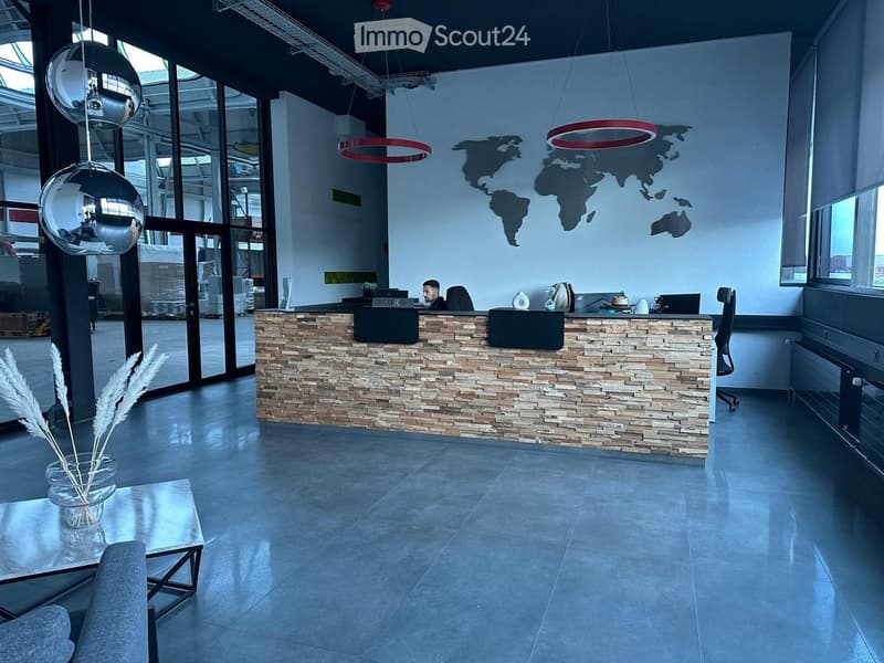 Modern reception area with a stone-textured reception desk, a vase with dried plants on a small table, and a hanging globe lamp. A person is seated behind the desk with a laptop and office supplies.