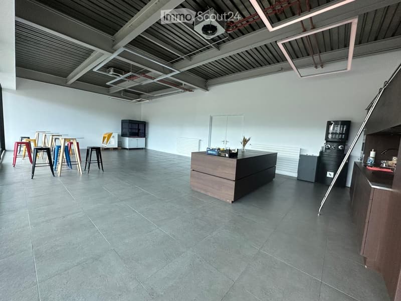Open-plan space with grey tiled floor, exposed metal ceiling beams, and industrial-style lighting. Features a central counter, stools, cabinets, and a sink area.