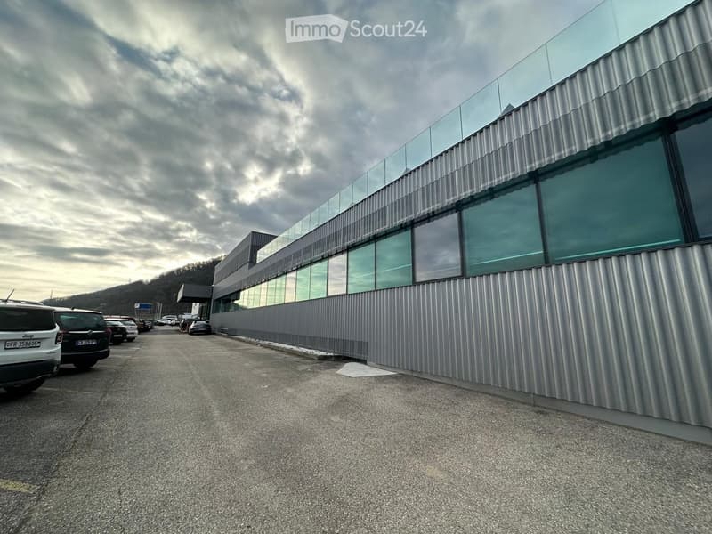 Modern building with a corrugated metal facade and large glass windows, surrounded by a parking area with several cars.