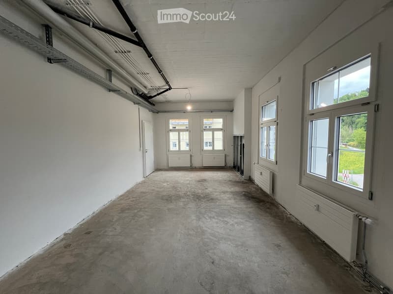 Empty room with bare concrete floor, white walls, multiple windows, and exposed pipes on the ceiling.