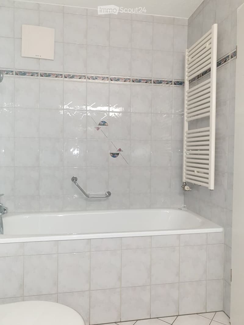 A clean, tiled bathroom with a white bathtub, a towel radiator, and a chrome grab bar for safety.